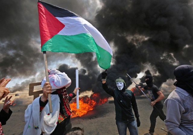 Un palestino sosteniendo su bandera nacional camina en el humo de neumáticos quemados junto a un manifestante con una máscara anónima durante enfrentamientos con las fuerzas israelíes en la frontera con la franja de Gaza al este de Khan Yunis el 14 de mayo de 2018, mientras los palestinos protestan por la inauguración de la embajada de los Estados Unidos después de su controvertido traslado a Jerusalén. / AFP PHOTO / SAID KHATIB